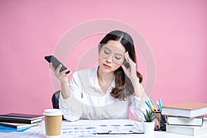 Young Asian woman wearing eyeglasses and a white shirt sits at a white desk with a laptop. She uses her smartphone to type sms