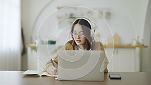 Young asian woman wearing glasses and headset working on computer laptop at house