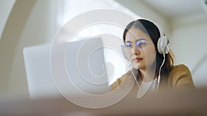 Young asian woman wearing glasses and headset working on computer laptop at house