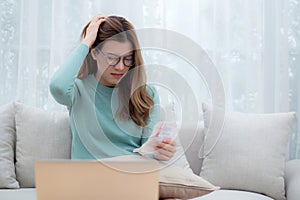 Young asian woman wearing glasses sitting on sofa worried with bills about financial and stress at home.