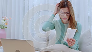 Young asian woman wearing glasses sitting on sofa worried with bills about financial and stress at home.