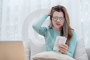 Young asian woman wearing glasses sitting on sofa worried with bills about financial and stress at home.