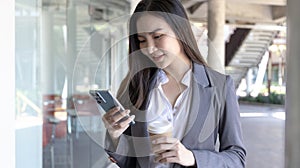 Young Asian woman wearing a gray suit is holding a phone and a paper coffee mug