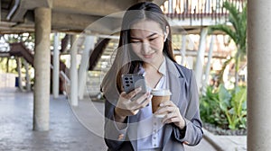 Young Asian woman wearing a gray suit is holding a phone and a paper coffee mug