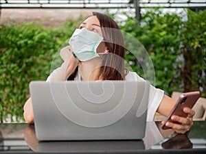 Young Asian woman wearing green medical mask looking up thinking while working with computer laptop and mobile phone at home