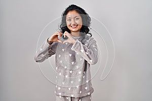 Young asian woman wearing pajama smiling in love doing heart symbol shape with hands