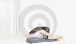 Beside of young asian woman wearing white undershirt and gray pants,exercising yoga while sitting on a white bed , stretch oneself