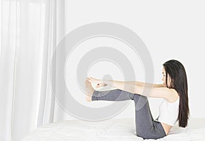 Beside of young asian woman wearing white undershirt and gray pants,exercising yoga while sitting on a white bed , stretch oneself