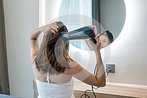 Young Asian woman in white dress sitting in front of mirror in hotel room and blow dry her wet hair after washing in the morning.