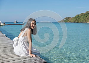 Young asian woman in white dress sitting on wooden pier in tropical sea on sunny day