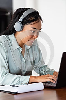 Young Asian woman working from home. Sitting in front of laptop computer with headphones, having online meeting