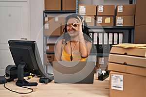 Young asian woman working at small business ecommerce wearing headset smiling happy doing ok sign with hand on eye looking through