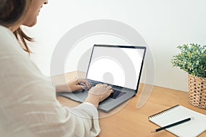 Young Asian woman working using and typing on laptop with mock up blank white screen while at home in office work space.