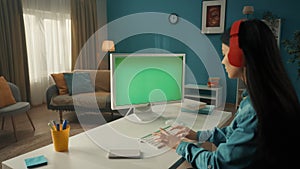 A young Asian woman works at a computer with a green screen close up. A longhaired brunette woman sits at a table in red