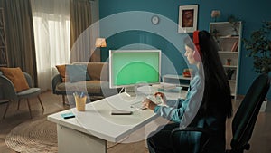 A young Asian woman works at a computer with a green screen. A longhaired brunette woman sits at a table in red wireless