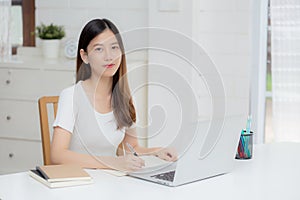 Young asian woman writing on notebook for planning working and using laptop computer on desk at home.
