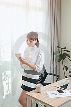 Young Asian woman writing notebook while standing behind working desk at home