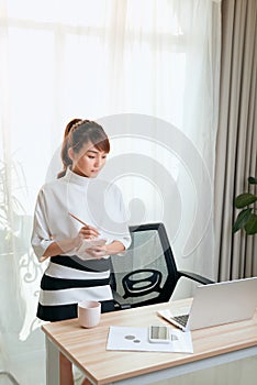 Young Asian woman writing notebook while standing behind working desk at home