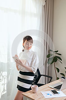 Young Asian woman writing notebook while standing behind working desk at home