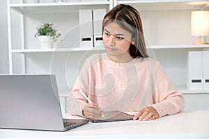 Young asian woman writting on digital tablet while looking at laptop computer, online learning, work from home