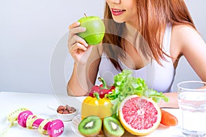 Young Asian women eating green apple with fruits, vegetables, water, dumbbell and tape measure on white table background.