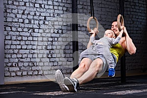 Young athlete father and little cute son exercising with gimnastic rings and smiling against brick wall in the gym.