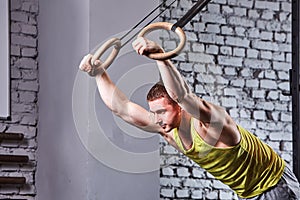 Young athlete man in the sportwear pulling up on gymnastic rings against brick wall in the cross fit gym.