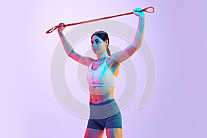 Young athlete woman in activewear doing exercises for hands using resistance band in neon light against gradient studio