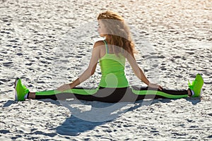 Young athletes - gymnast with curly hair, light green suit and sneakers doing the splits on the beach in summer, morning exercise.