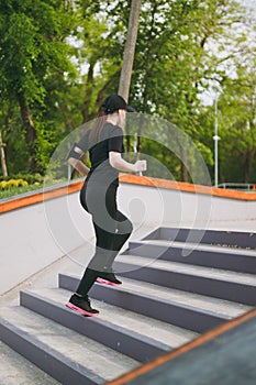 Young athletic beautiful brunette girl in black uniform, cap with headphones doing sport exercises, training and running