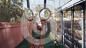 Young athletic boy doing exercise on the gymnastic rings at street workout place. Young athlete doing pull-ups on