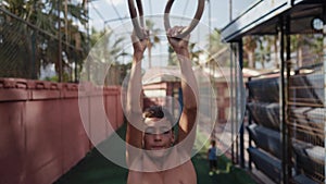 Young athletic boy doing exercise on the gymnastic rings at street workout place. Young athlete doing pull-ups on