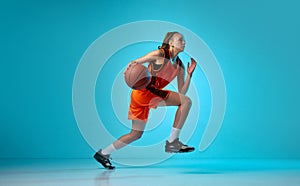 Young athletic girl, basketball player in motion, running with ball against blue studio background in neon light