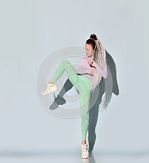 Young athletic girl with dreadlock hairstyle in tight sportswear stands holding knee up making kick by leg