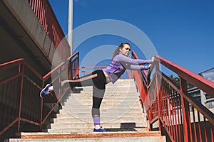 Young athletic girl stands with her foot on the railing of the stairs, workout urban sport concept