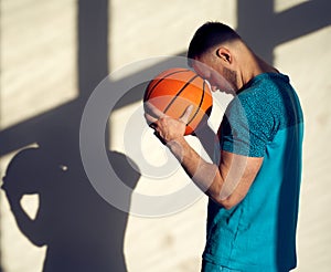 Young athletic man, basketball player holding ball and standing near wall with shadows from window