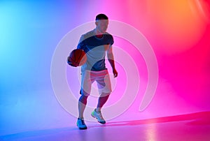Young athletic man dribbling with basketball ball posing on mix of blue and pink background with light projection