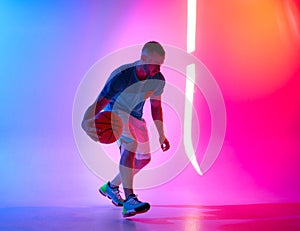 Young athletic man dribbling with basketball ball posing on mix of blue and pink background with light projection