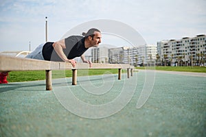 Young athletic man in sportswear, doing bar push-ups on crossbar, exercising outdoors in the urban sportsground.