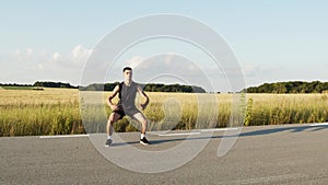 Young athletic man in sportwear doing squats with jumps on path at summer nature