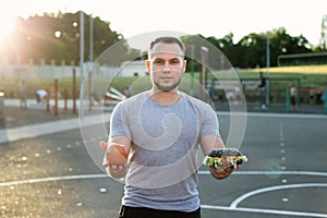 Young athletic man in a t-shirt holds a Burger and an Apple in the background of the stadium, a choice of healthy and