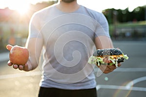 Young athletic man in a t-shirt holds a Burger and an Apple in the background of the stadium, a choice of healthy and