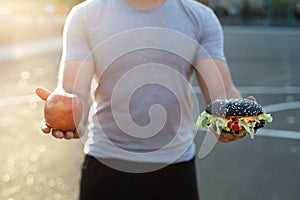 Young athletic man in a t-shirt holds a Burger and an Apple in the background of the stadium