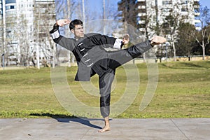 Young athletic martial arts fighter practicing kicks in a public park wearing kung fu uniform