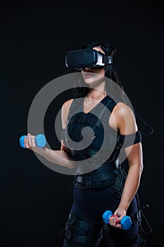 A young athletic woman in an EMS suit and virtual reality glasses with dumbbells on an isolated black background. EMS