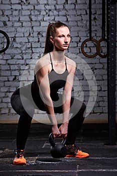 Young athletic woman exercising with kettlebell while being in squat position against brick wall.