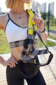 Young athletic woman with perfect body in sportswear holds in hands trx fitness straps for training in the city park