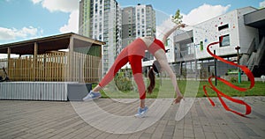 Young athletic woman in red performs cartwheel with gymnastic ribbon in the yard of an apartment building, gymnastics in