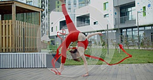 Young athletic woman in red performs cartwheel with gymnastic ribbon in the yard of an apartment building, gymnastics in