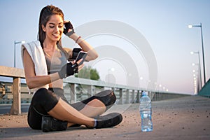 Young athletic woman using phone while siting on sidewalk and resting after running over the bridge.